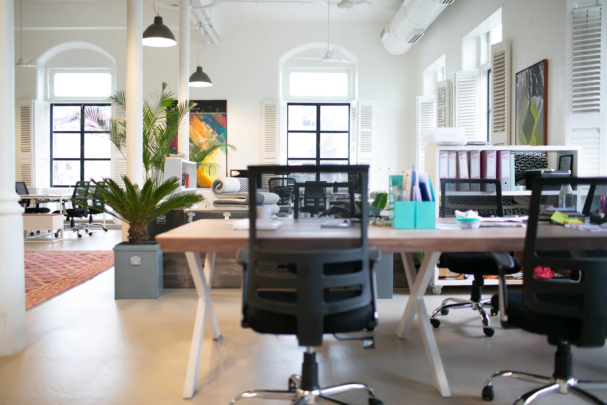 An open-plan office floor with rows of wooden desks, task chairs and tall windows